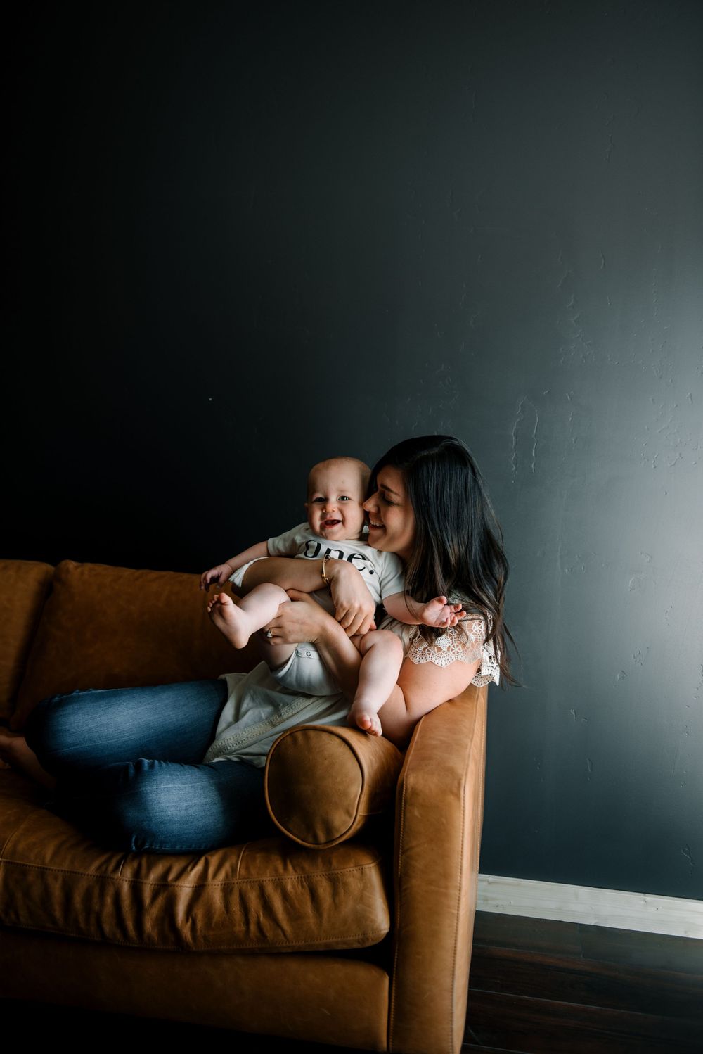 Parent and infant share joyful moment on leather couch with dark wall backdrop.