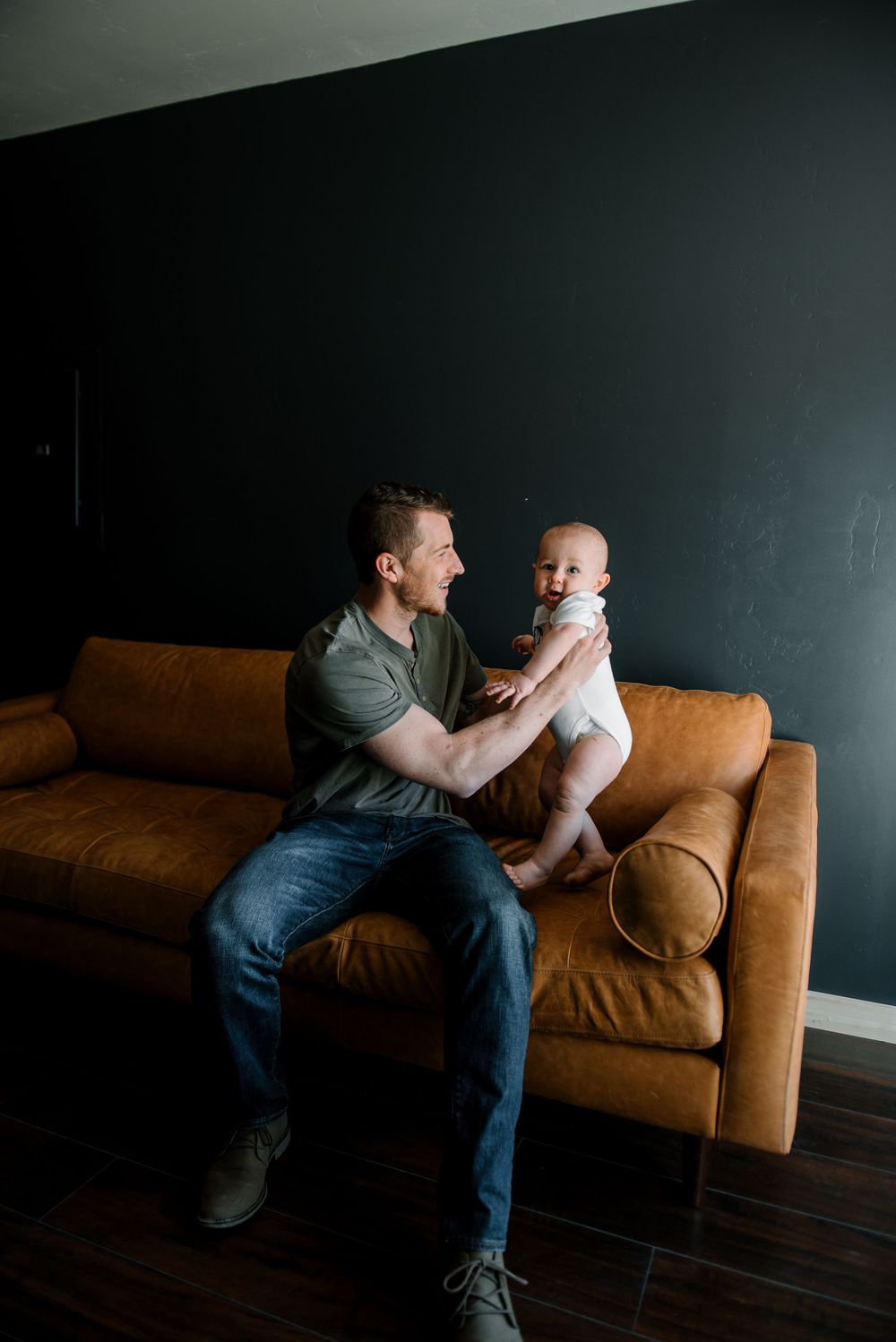 Father and baby share playful interaction on tan leather sofa in moody setting.