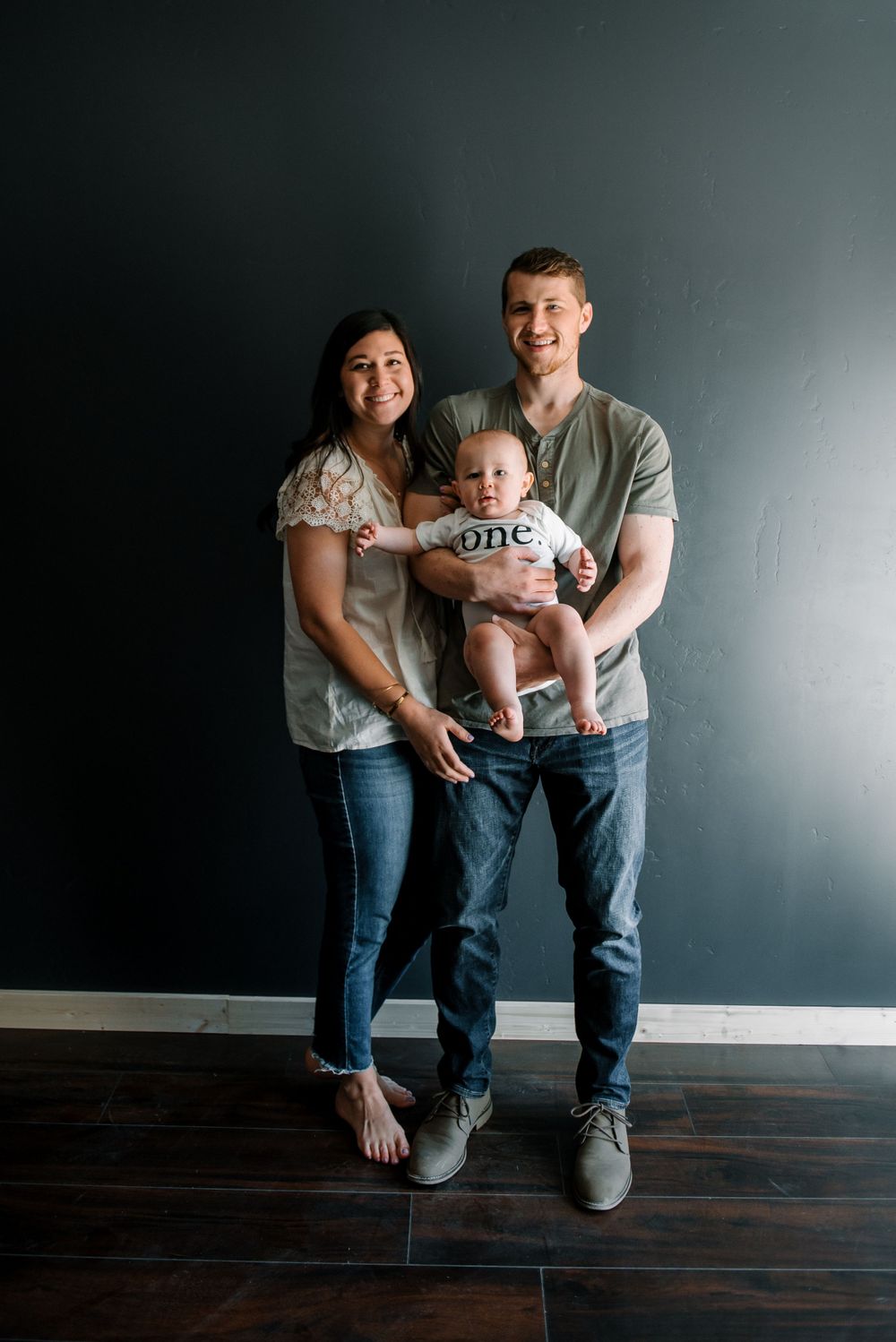 Family portrait against dark wall with baby wearing 'one' onesie.