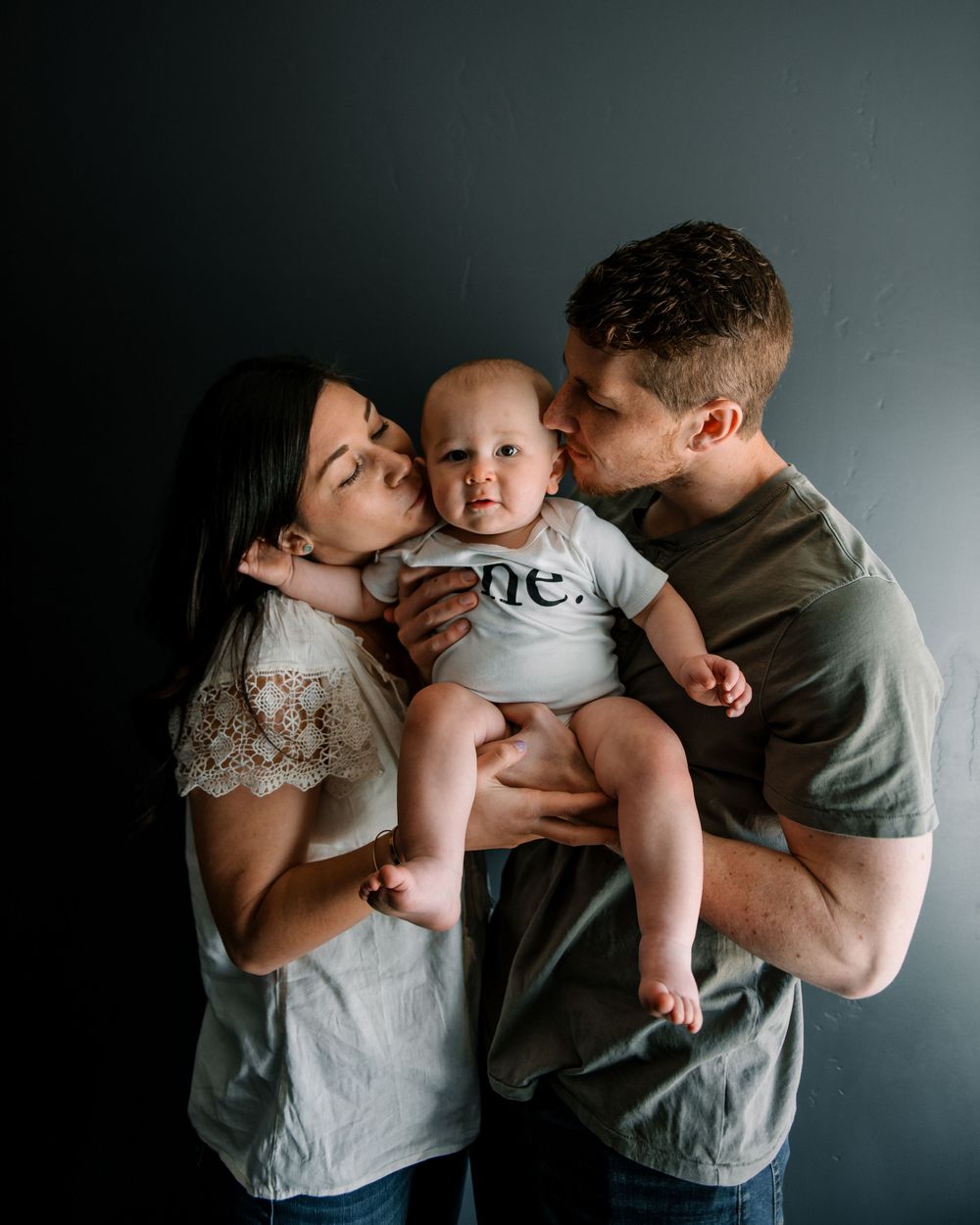 Intimate family portrait with baby in 'one' onesie against dark background.