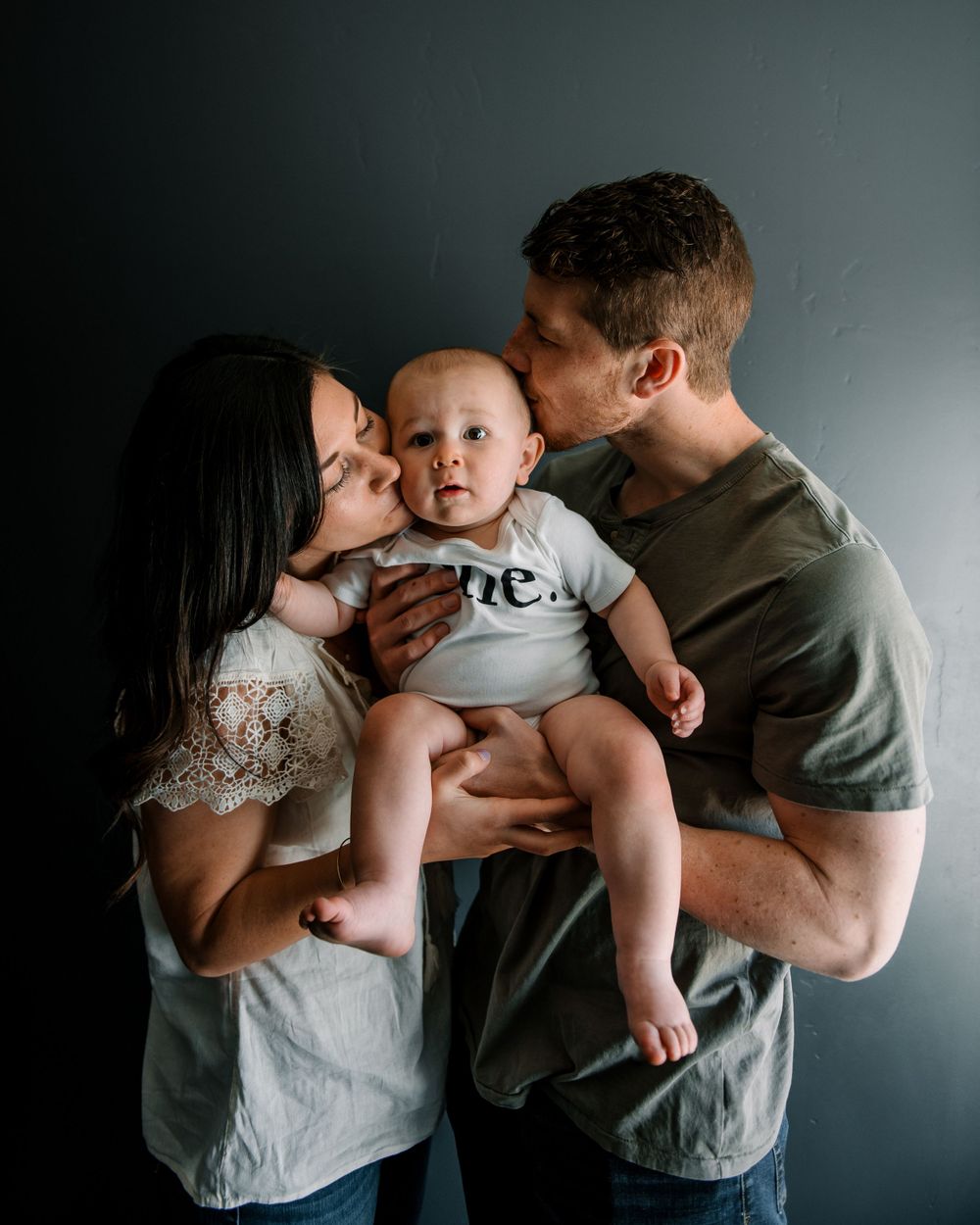 Parents kissing baby in 'one' onesie during intimate photo session.