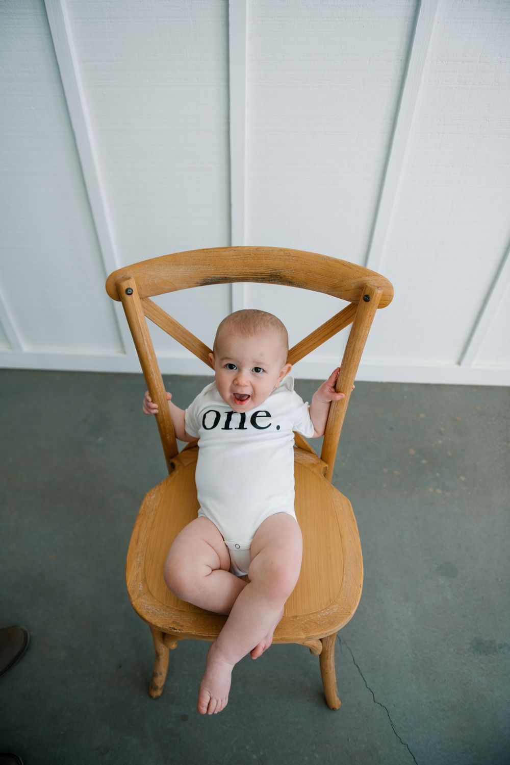 Baby in white 'one' onesie sits in wooden chair against white wall.