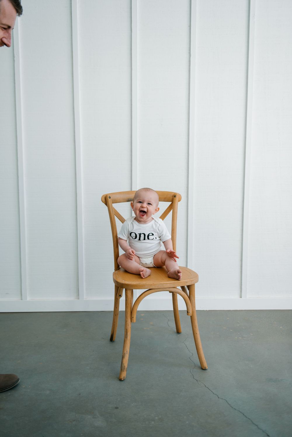 Happy baby in 'one' onesie laughs while sitting on wooden chair.