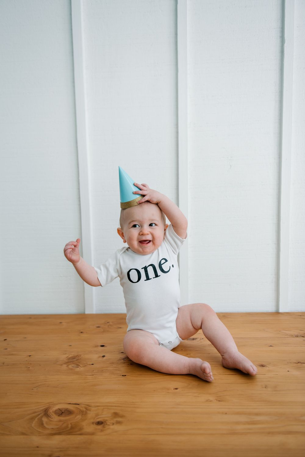 Baby wearing party hat and 'one' onesie sits on wooden floor laughing.