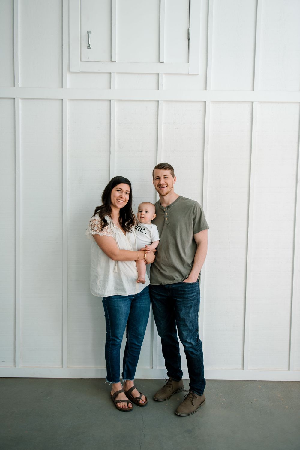 A family poses together against a white paneled wall wearing casual clothing.