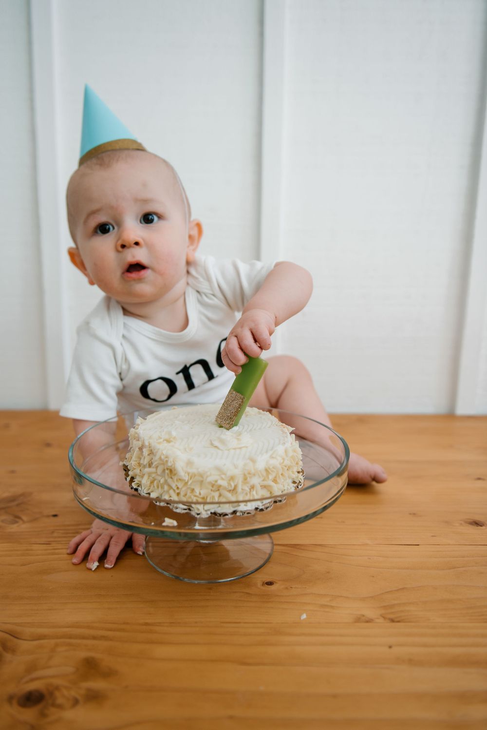 Baby wearing 'one' shirt and party hat reaches for birthday cake on glass stand.