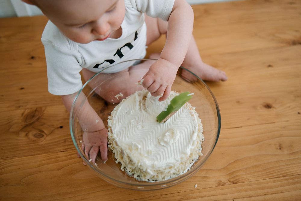 Baby in white shirt leaning over a white frosted birthday cake on wooden table.