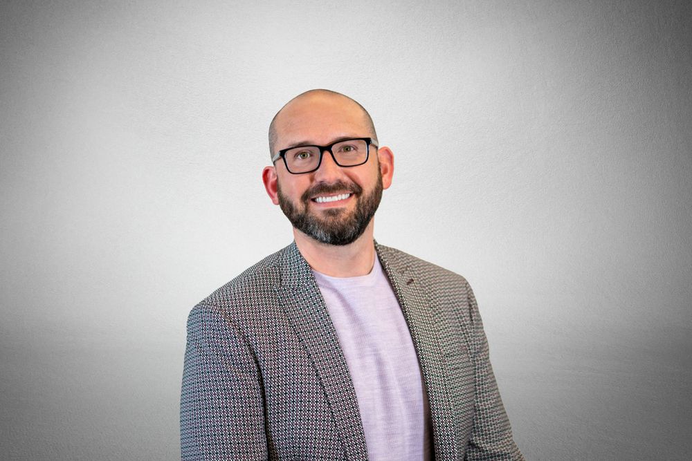 Professional wearing glasses and a plaid blazer over a lilac shirt poses against a gray backdrop. Quad Cities Business Headshot Photography