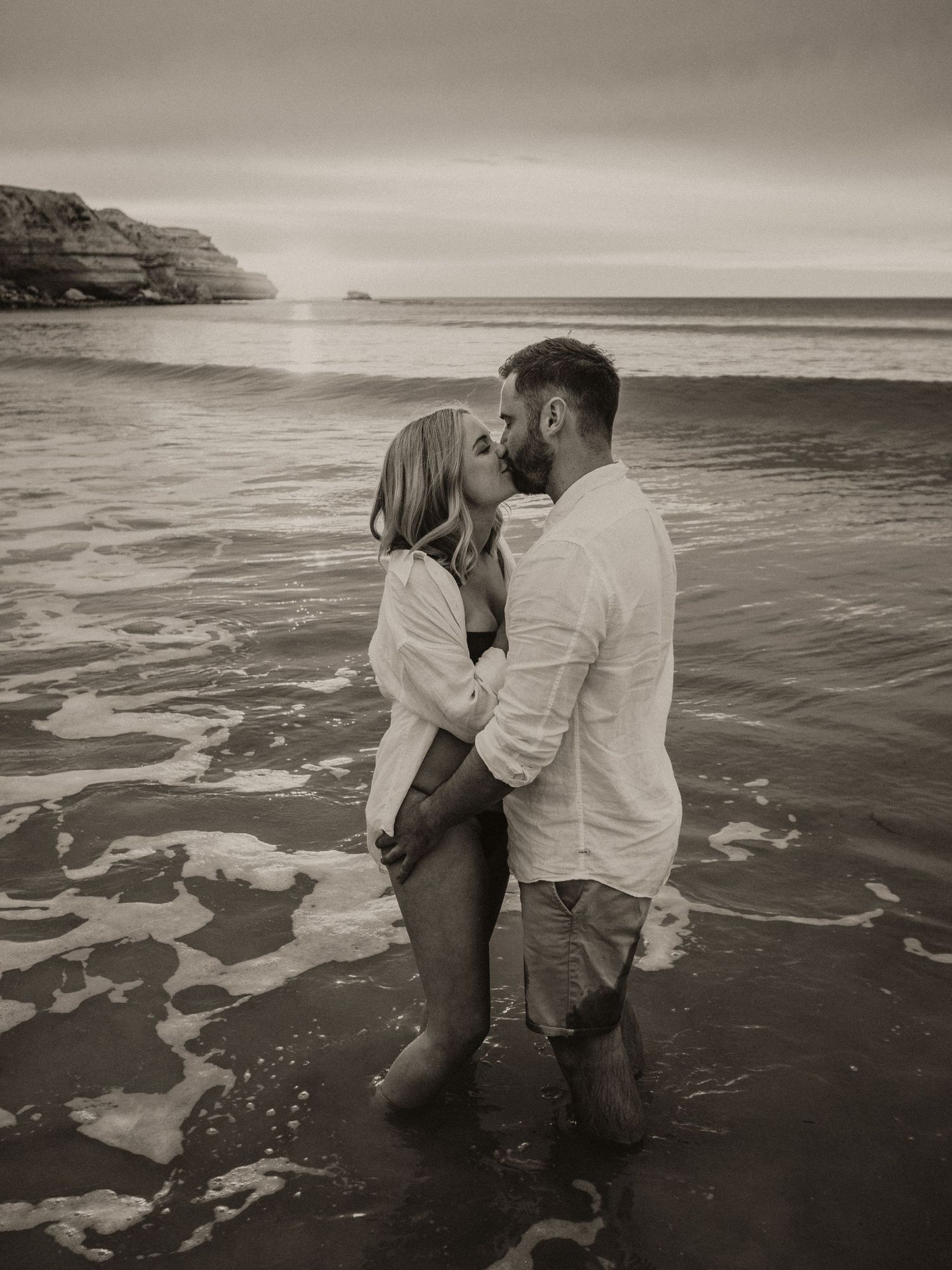 Black and white photo of couple standing in ocean waves at sunset.
