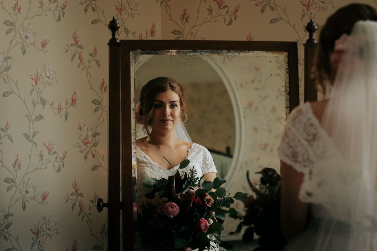 Bride in vintage-style dress glances at a mirror during wedding preparations in a floral wallpapered room.