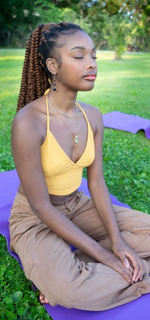 Person in yellow top and beige pants relaxing on purple yoga mat outdoors.