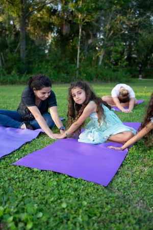 Three yoga practitioners stretching together on purple mats in a grassy outdoor setting during sunset.