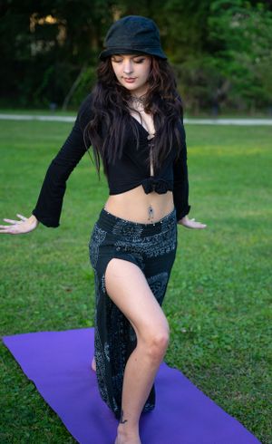 Dancer in black outfit and bucket hat performing movement on purple yoga mat outdoors.