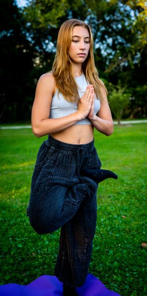 Yoga practitioner in grey crop top and dark pants performing tree pose on purple mat outdoors.