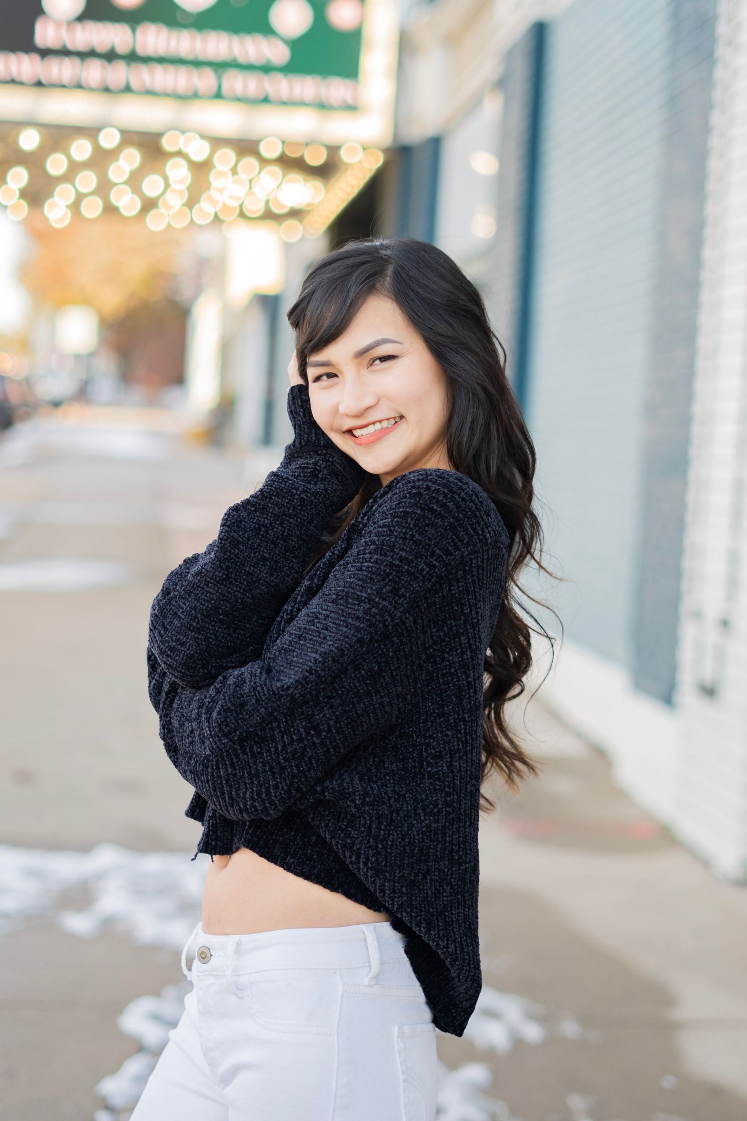 Smiling young woman in a black knit sweater and white jeans posing on a snowy city sidewalk.
