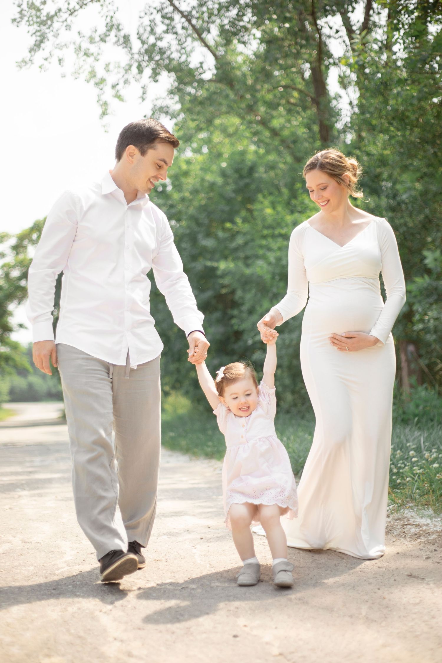 Sunlit outdoor family photo on a tree-lined path in white and neutral tones.