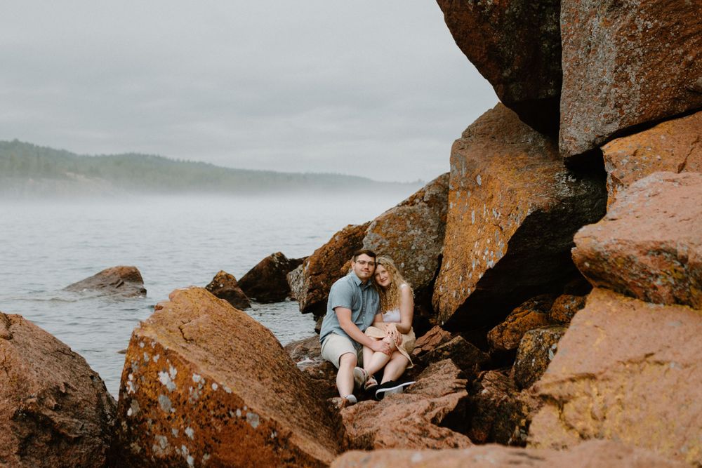 Couple sits together on rocky coastal outcrop overlooking misty ocean view.