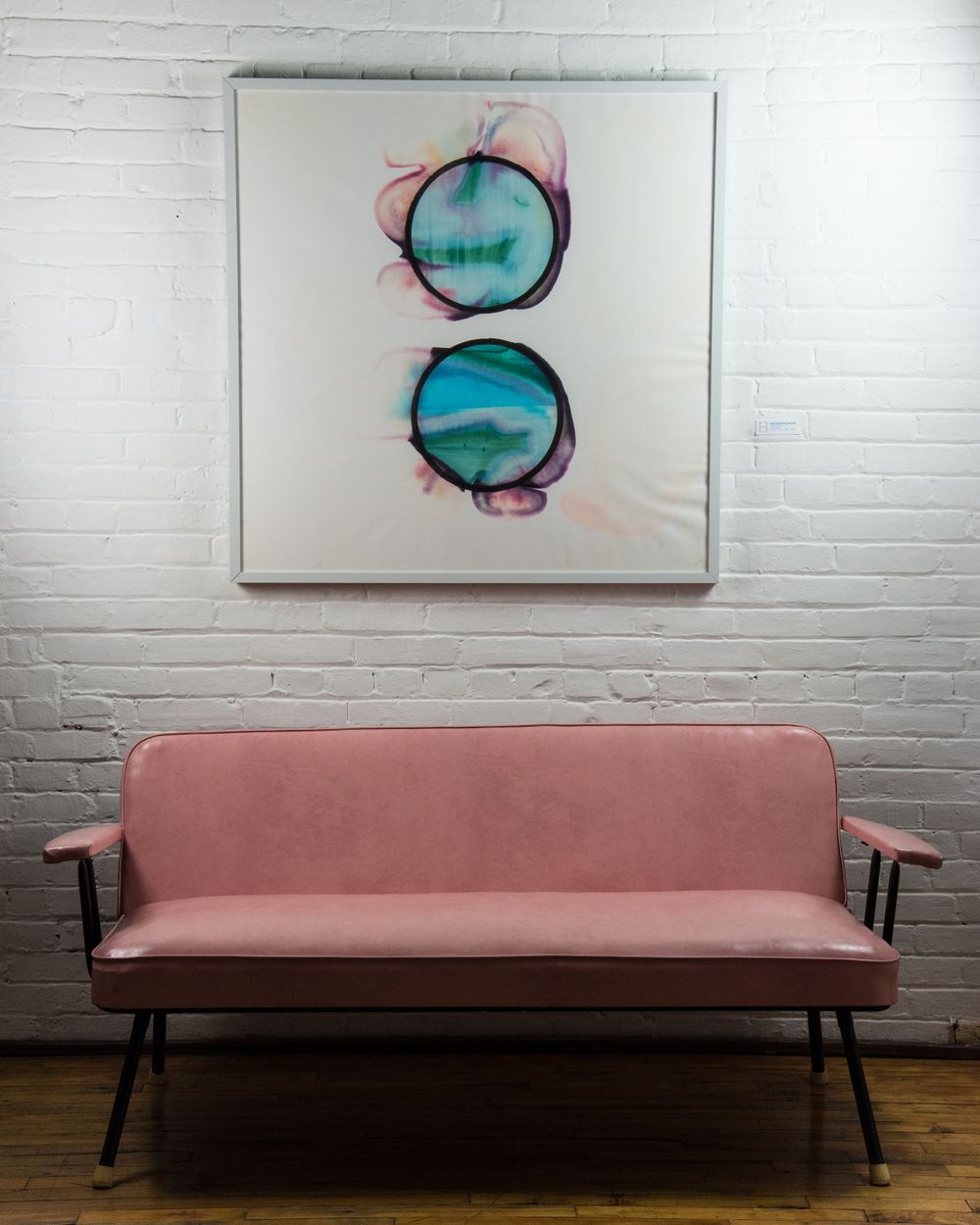 Minimalist turquoise circular artwork displayed above pink mid-century modern bench against white brick wall.
