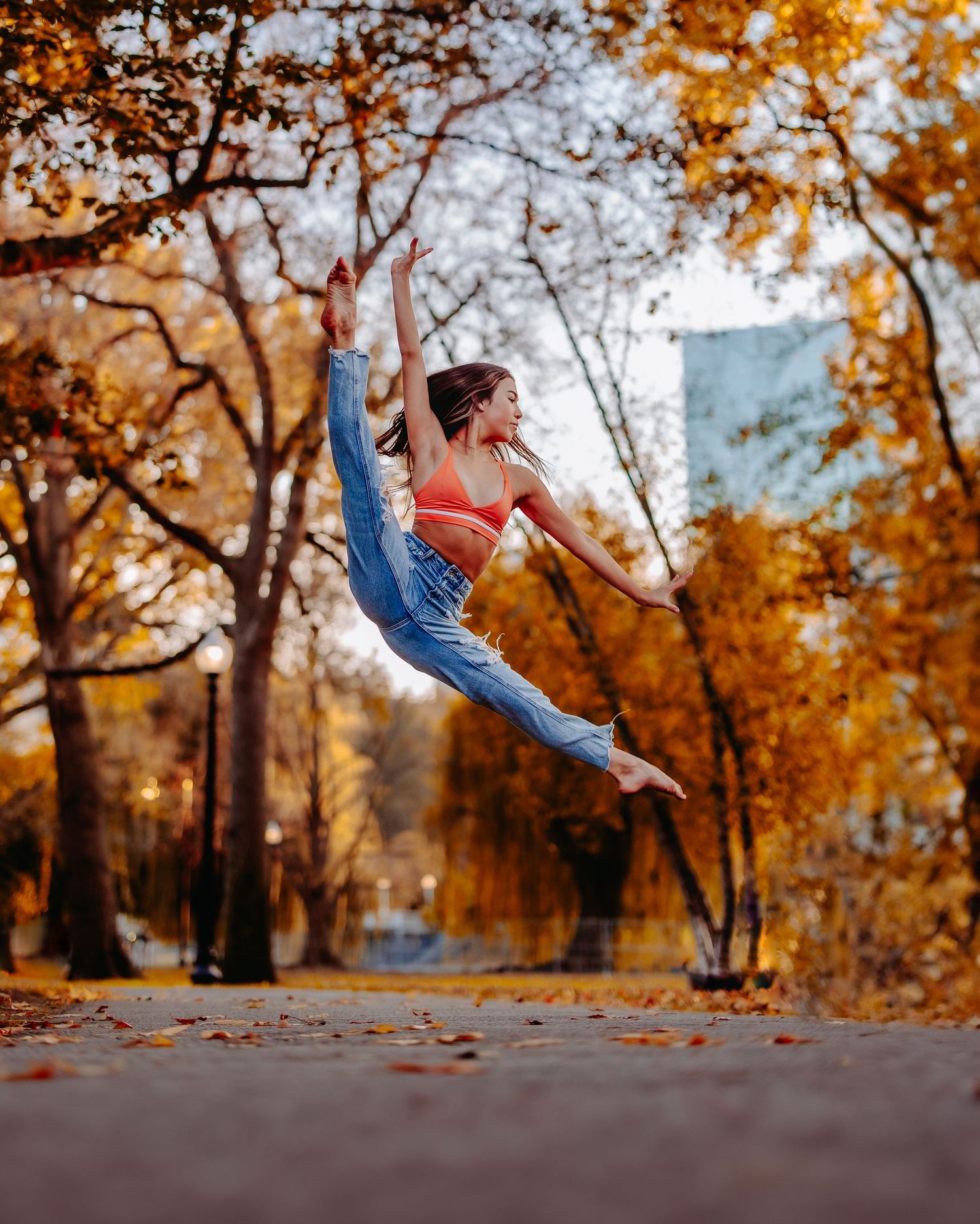 Dancer leaps gracefully through autumn trees in urban park with orange foliage.