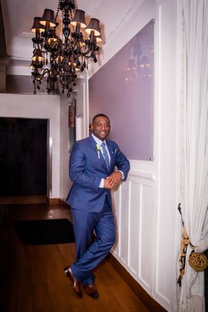 A groom in a blue suit poses against a white wall with elegant chandeliers.