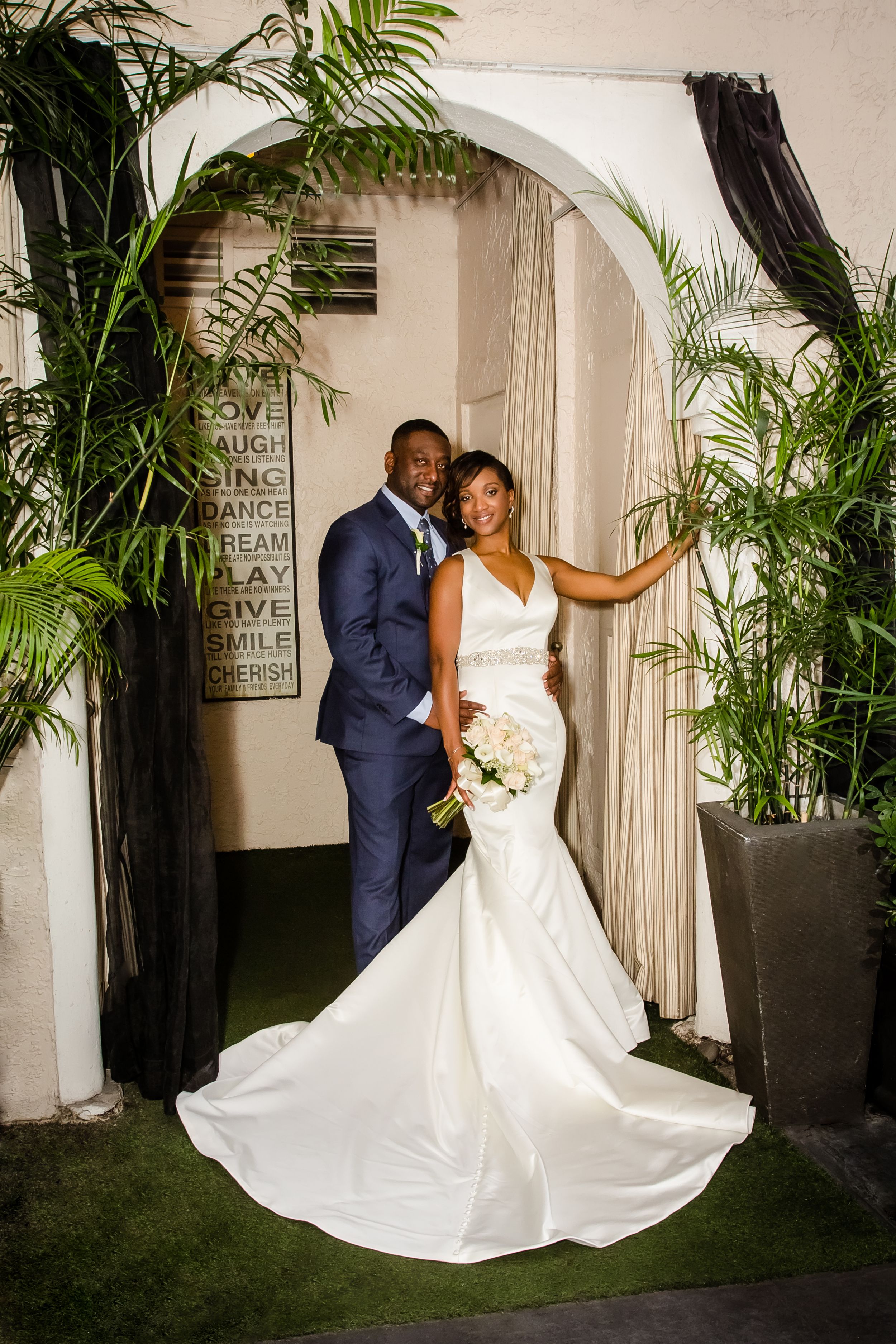 Newlyweds pose in an elegant garden setting with white columns and tropical plants creating a romantic backdrop.