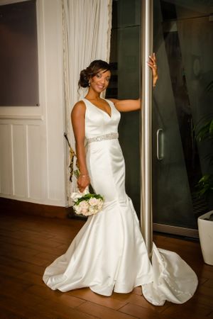 Elegant bride in white satin gown poses gracefully against interior wall with bouquet.
