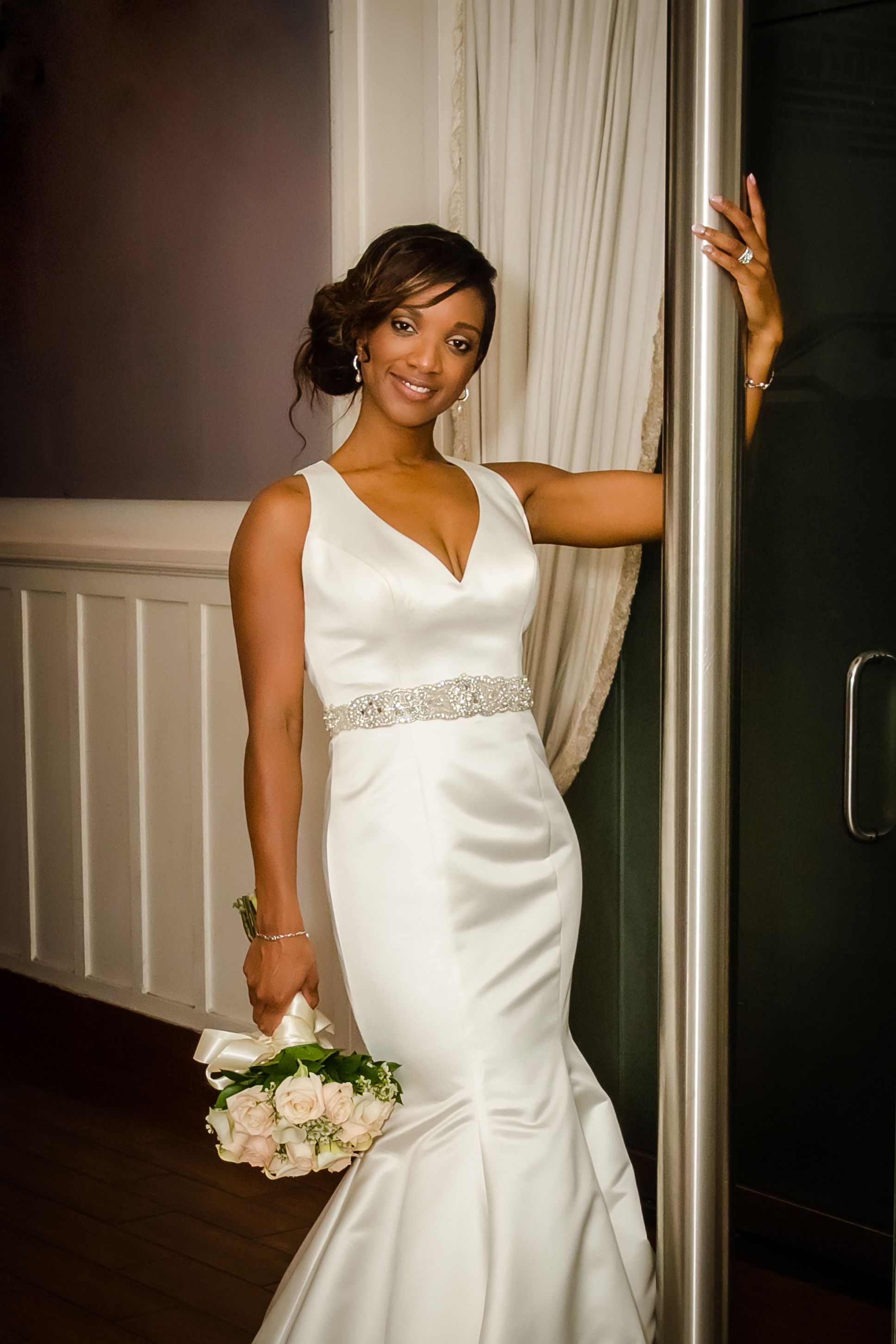 Bride in elegant white dress with beaded waistline poses against interior wall.