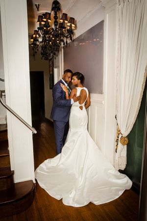 Newlyweds dance together in an elegant room with white curtains and dark chandeliers.