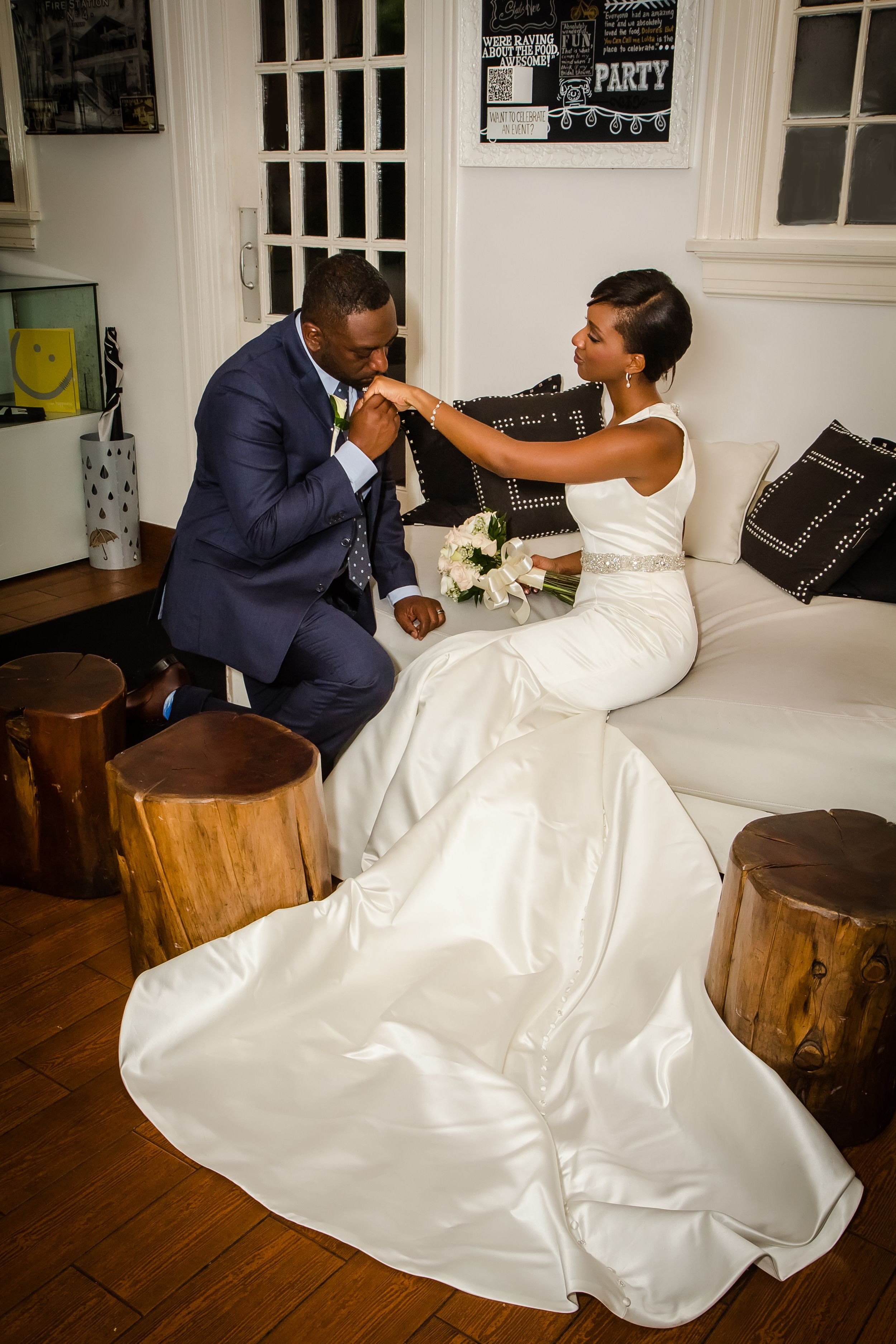 Bride in flowing white gown sits playfully with groom on rustic wooden furniture during wedding photoshoot.