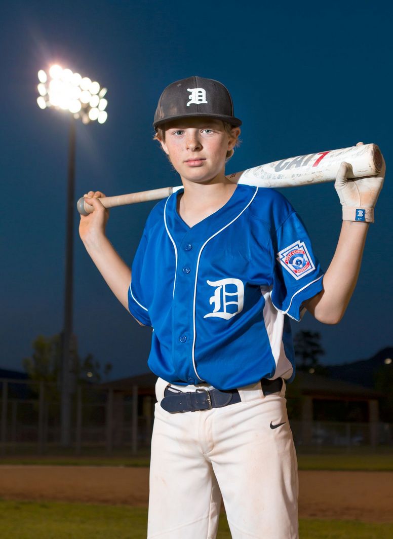 Young baseball player in Detroit Tigers uniform poses with bat under stadium lights.