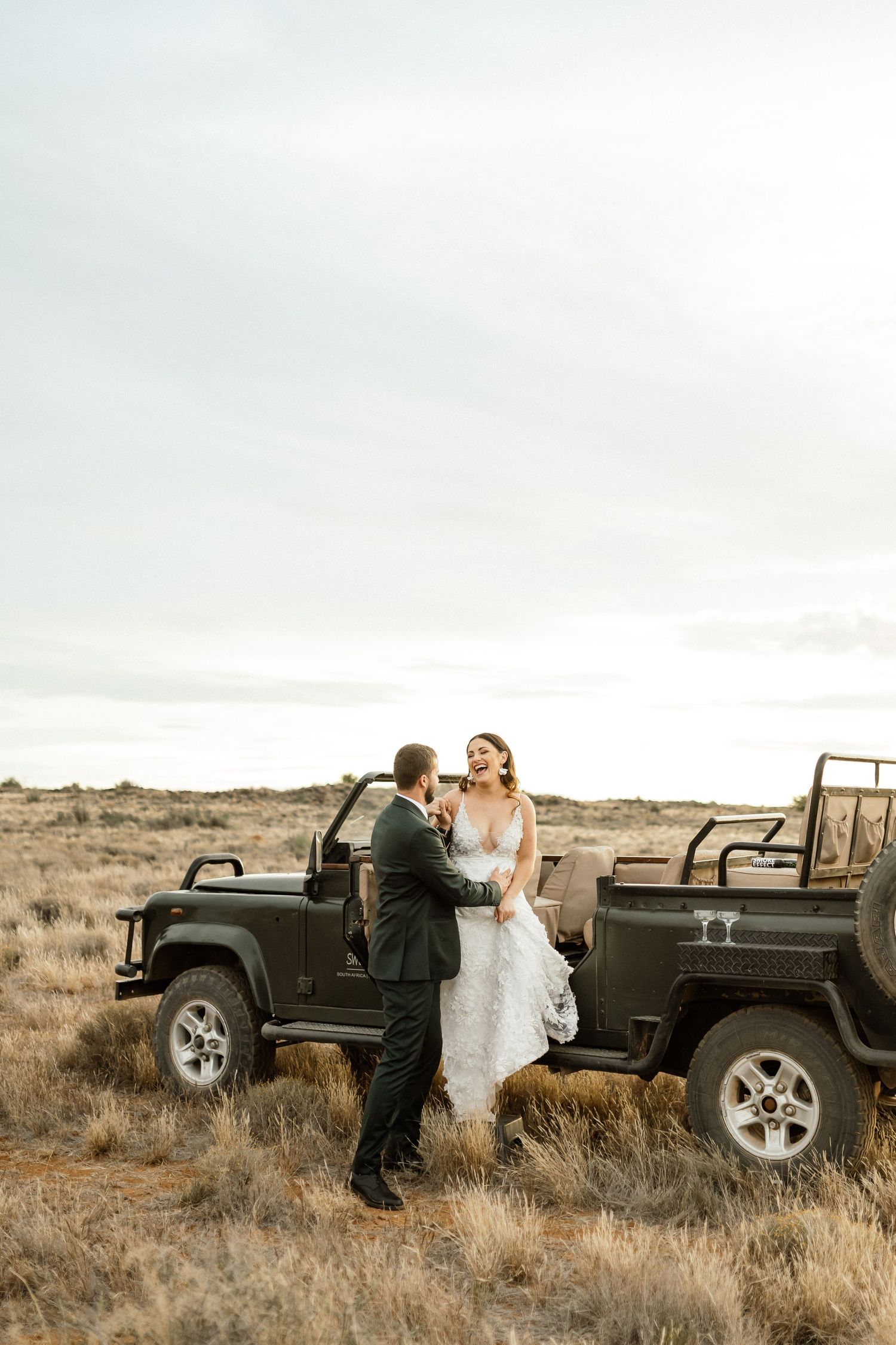 Laughing bride and groom sharing a moment on a Land Cruiser in a sunlit golden field