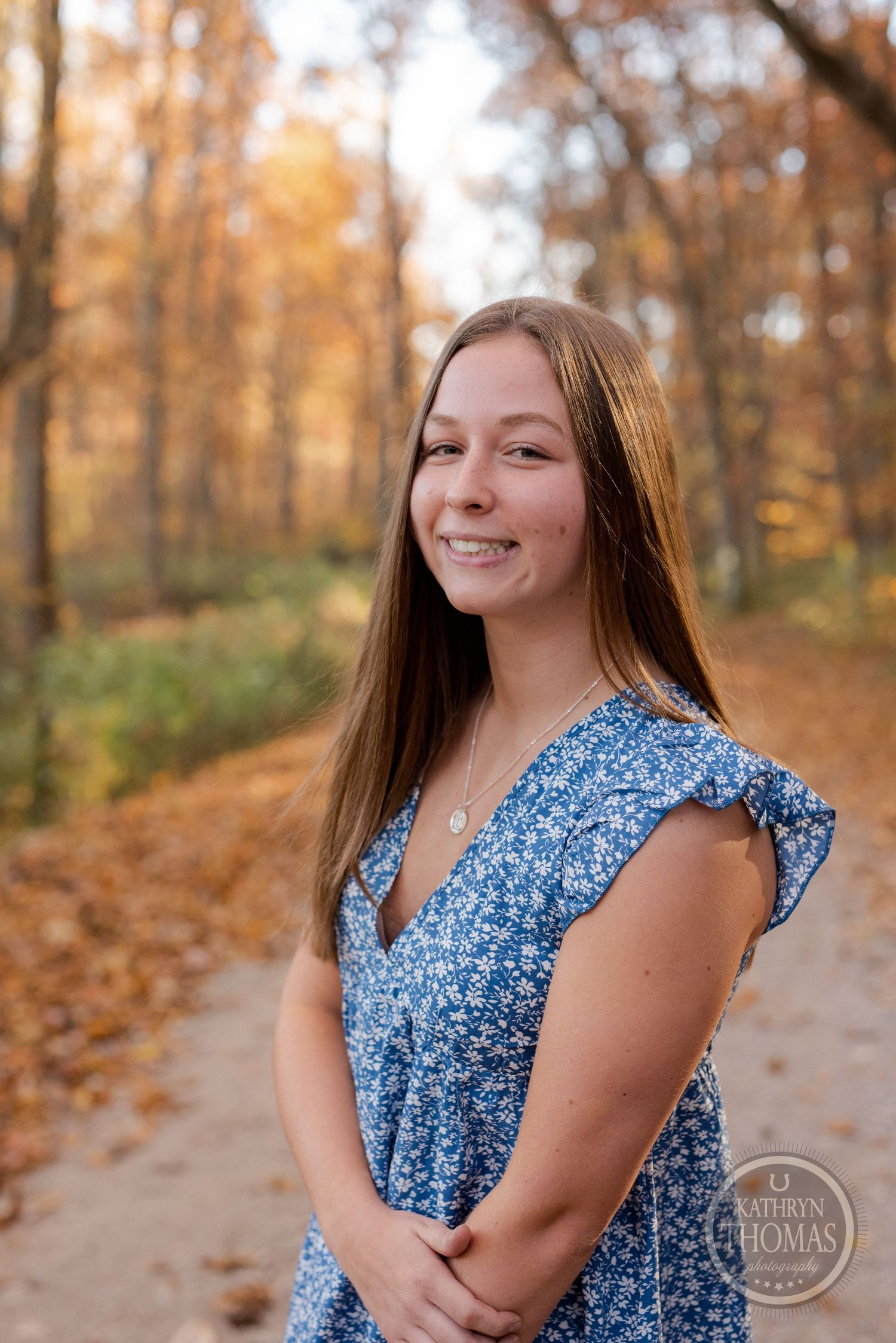 A person wearing a blue floral dress poses on a wooded path covered in fallen autumn leaves.