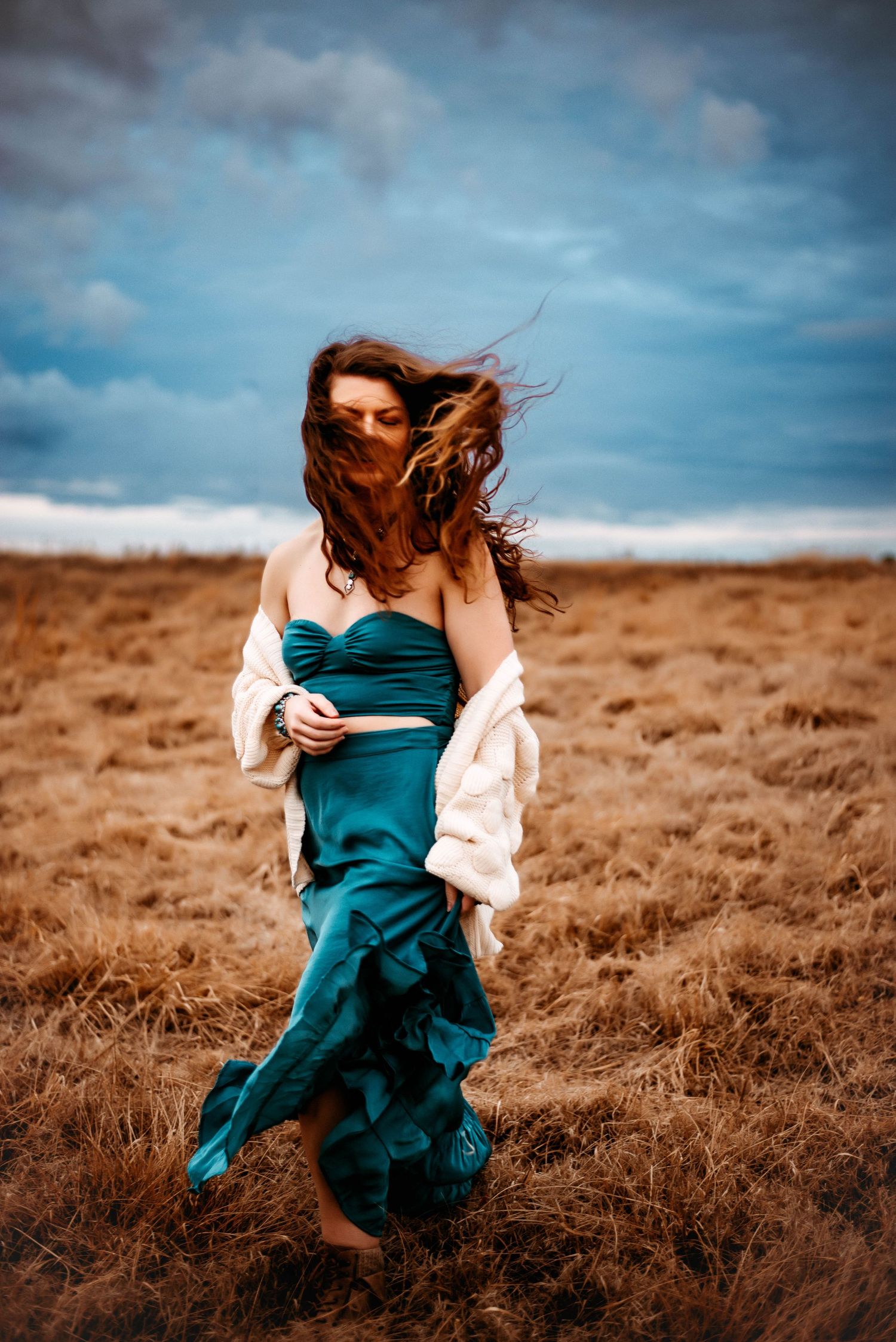 Figure in flowing teal dress dancing through a windswept field under moody skies.