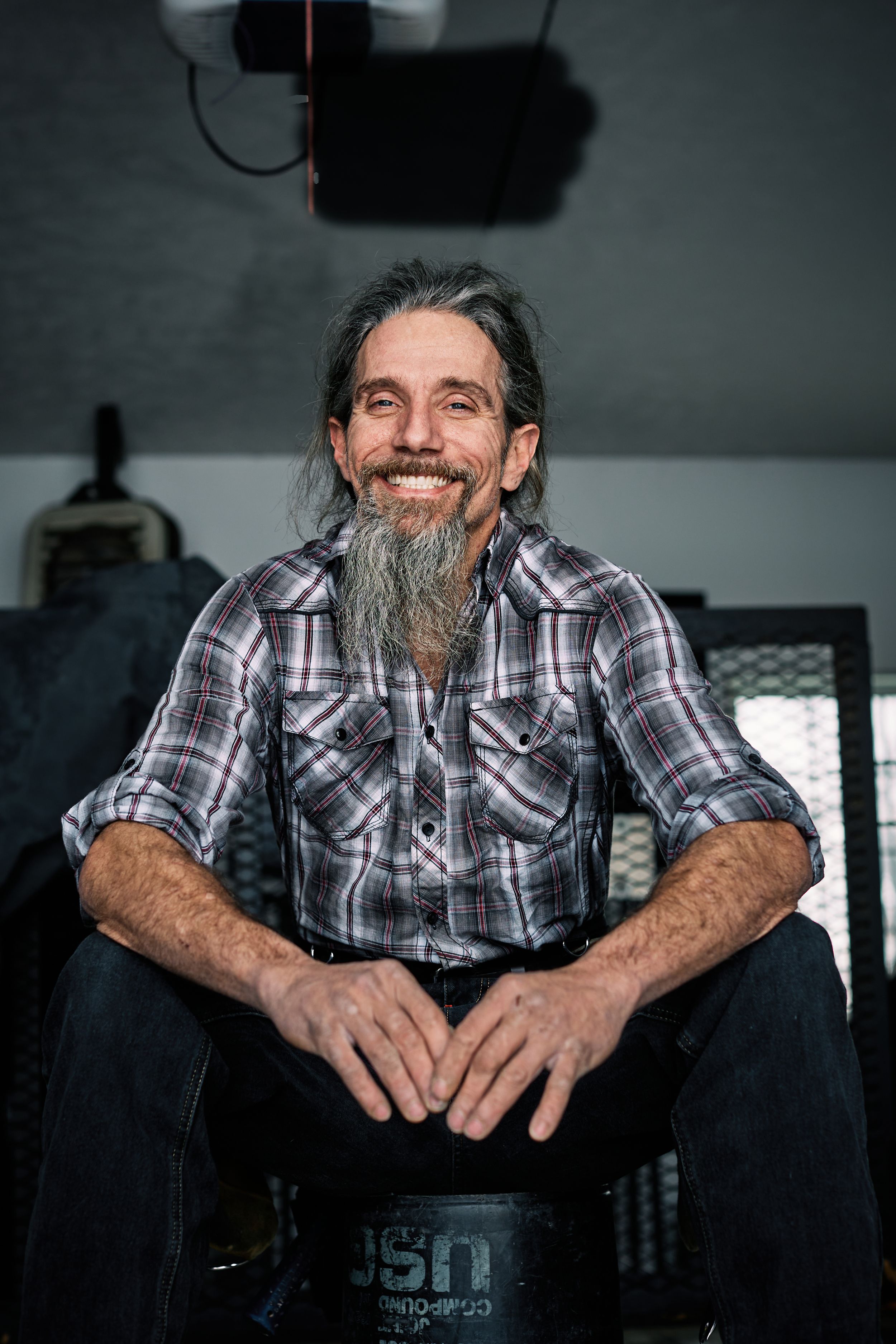 Craftsman with long beard sits casually in dimly lit workshop.
