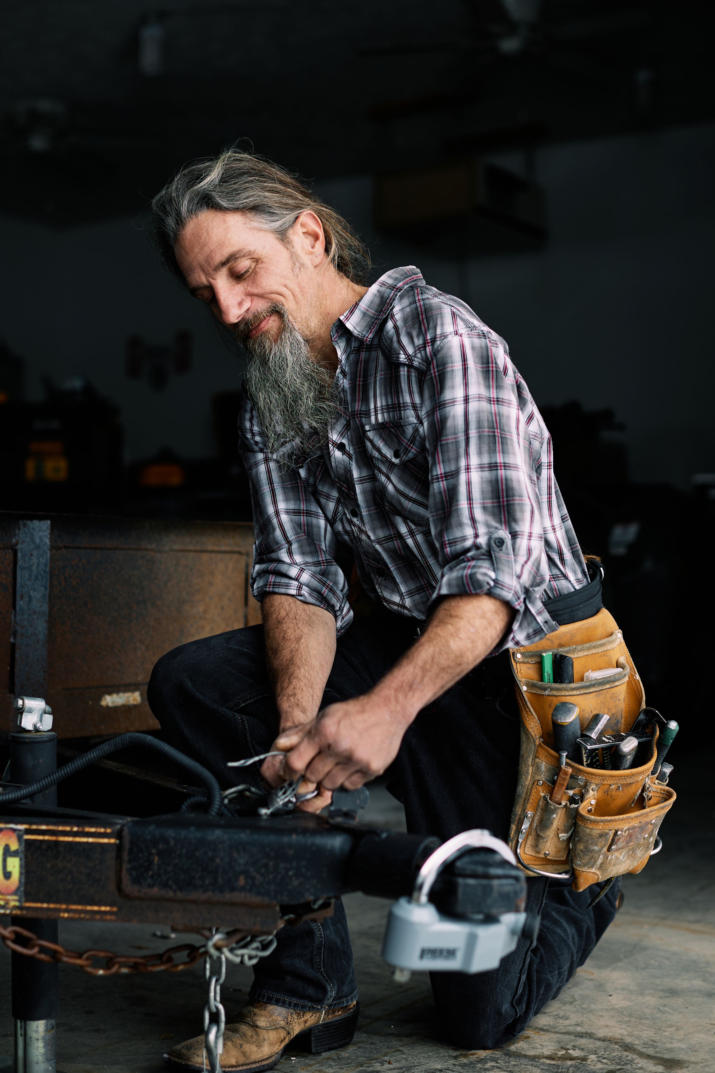 Skilled worker with tool belt kneels while working in dark workshop setting.