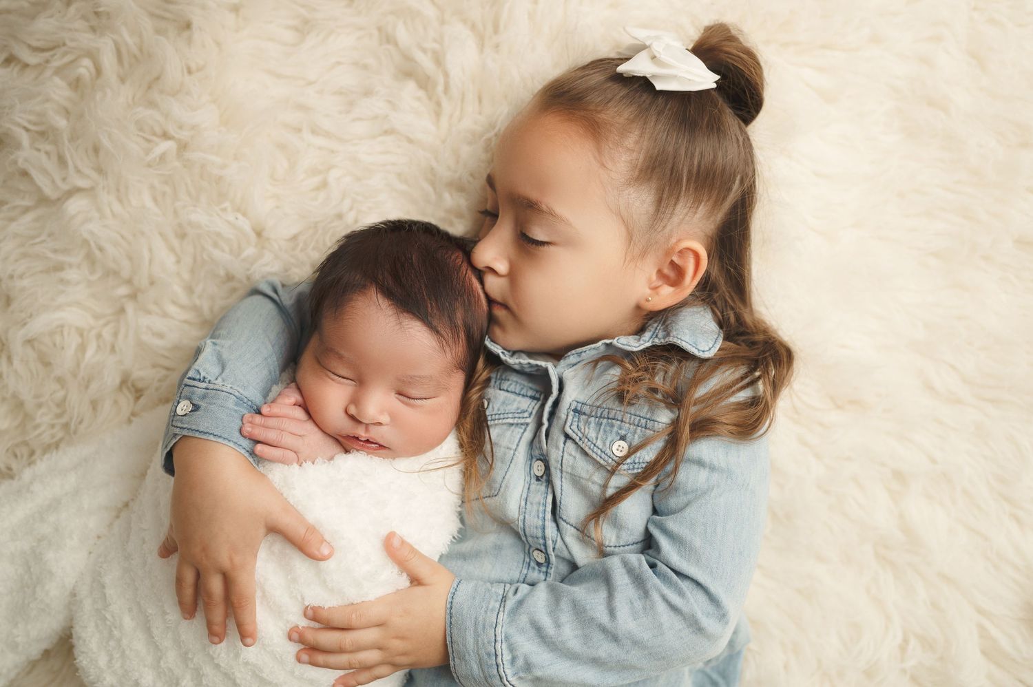 Young child in denim jacket gently kisses sleeping baby on soft cream blanket.