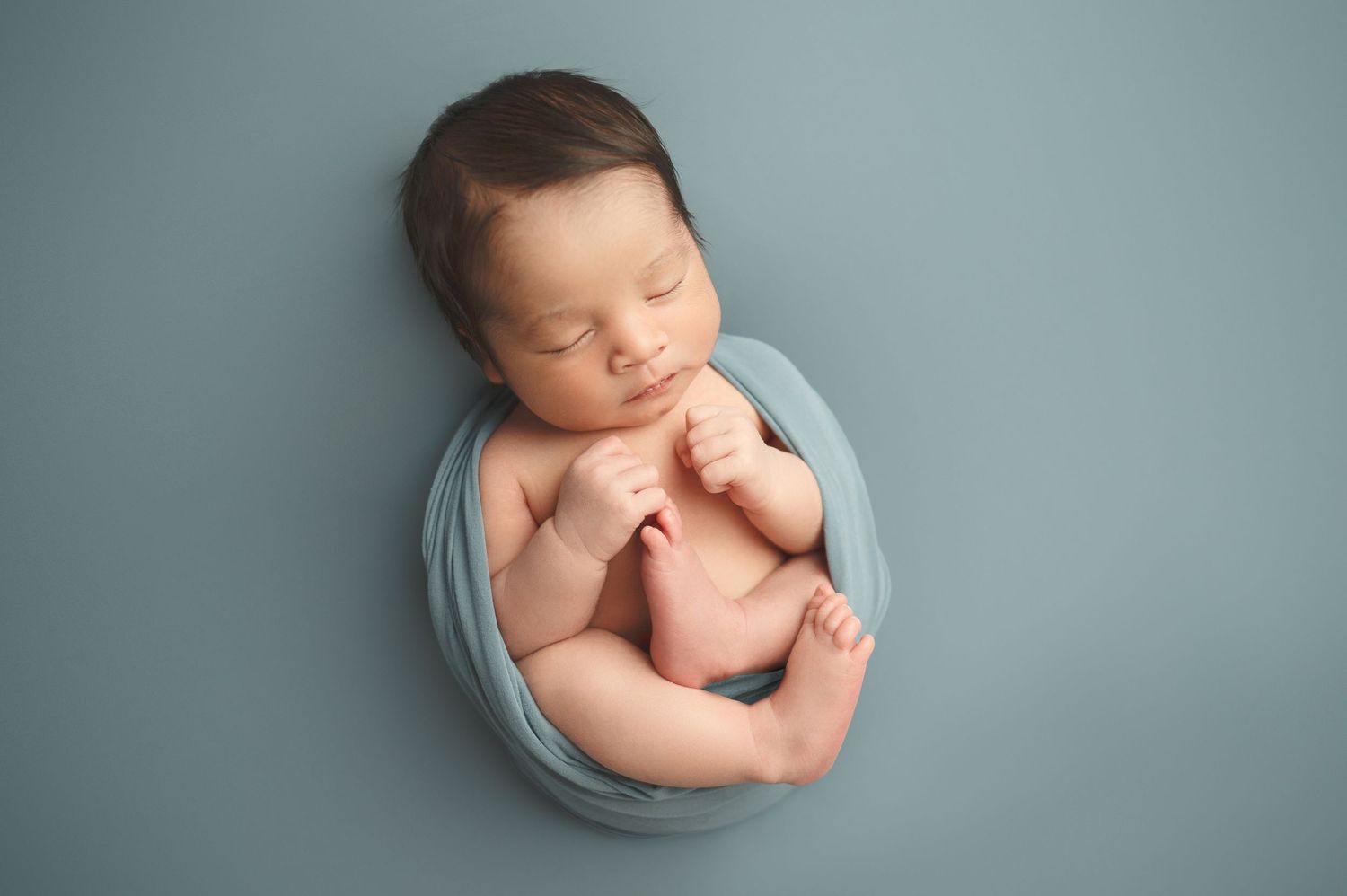 Newborn baby wrapped in soft blue fabric curled up in fetal position during professional photo session.
