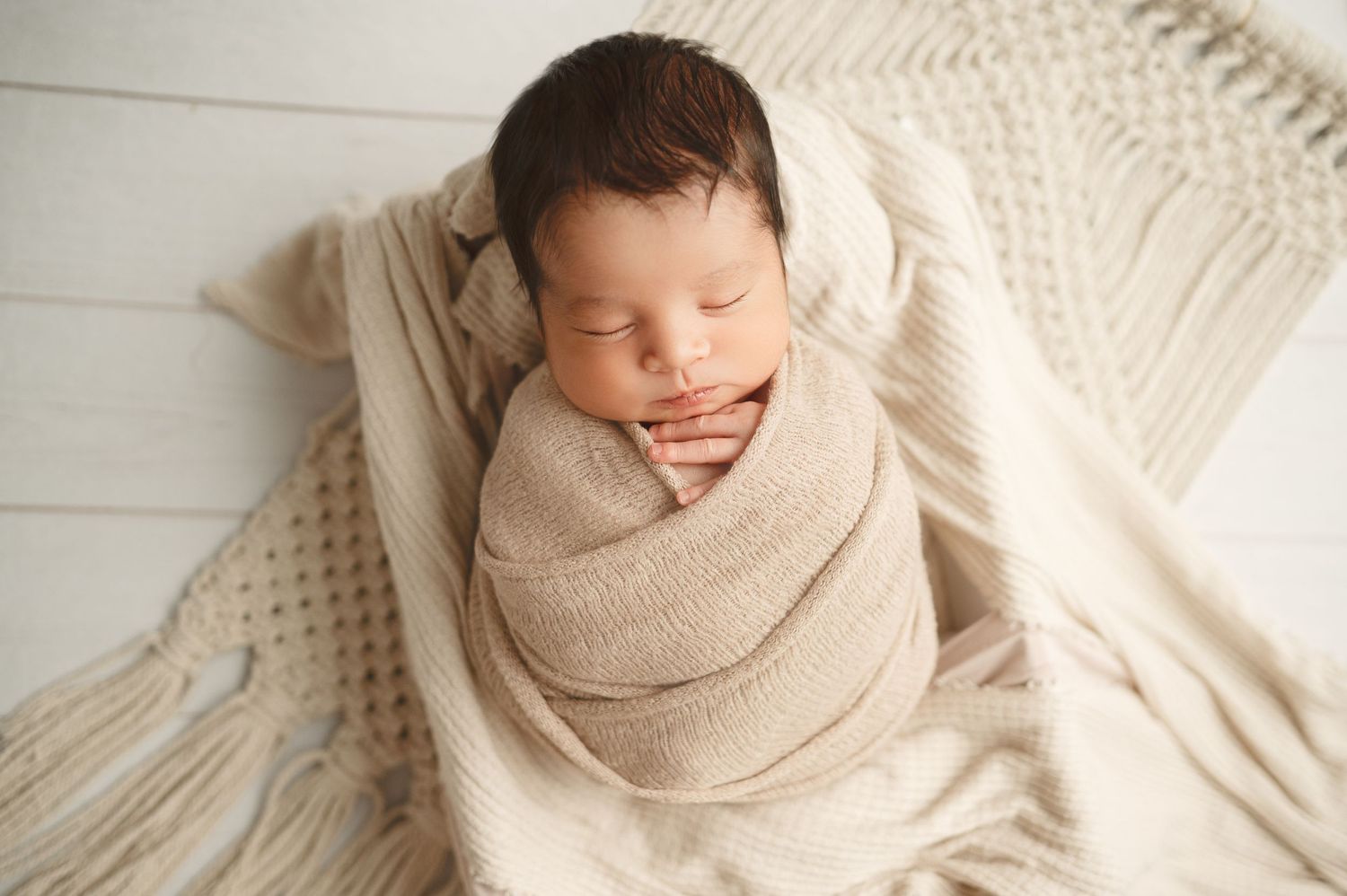 Newborn baby wrapped in beige knit blanket sleeping on white textured surface.