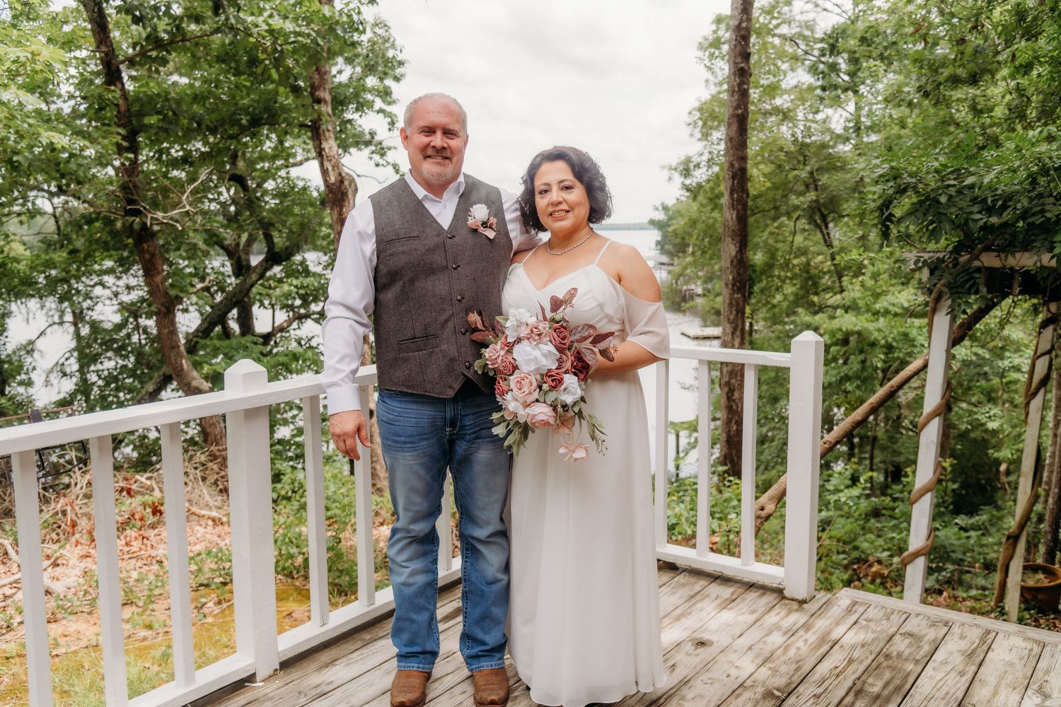 Couple on wooden deck during autumn dressed in casual wedding attire holding bouquet.