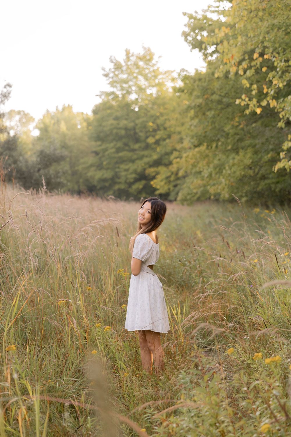 teenage girl in a grassy field in the morning smiling for a photo with a white dress on