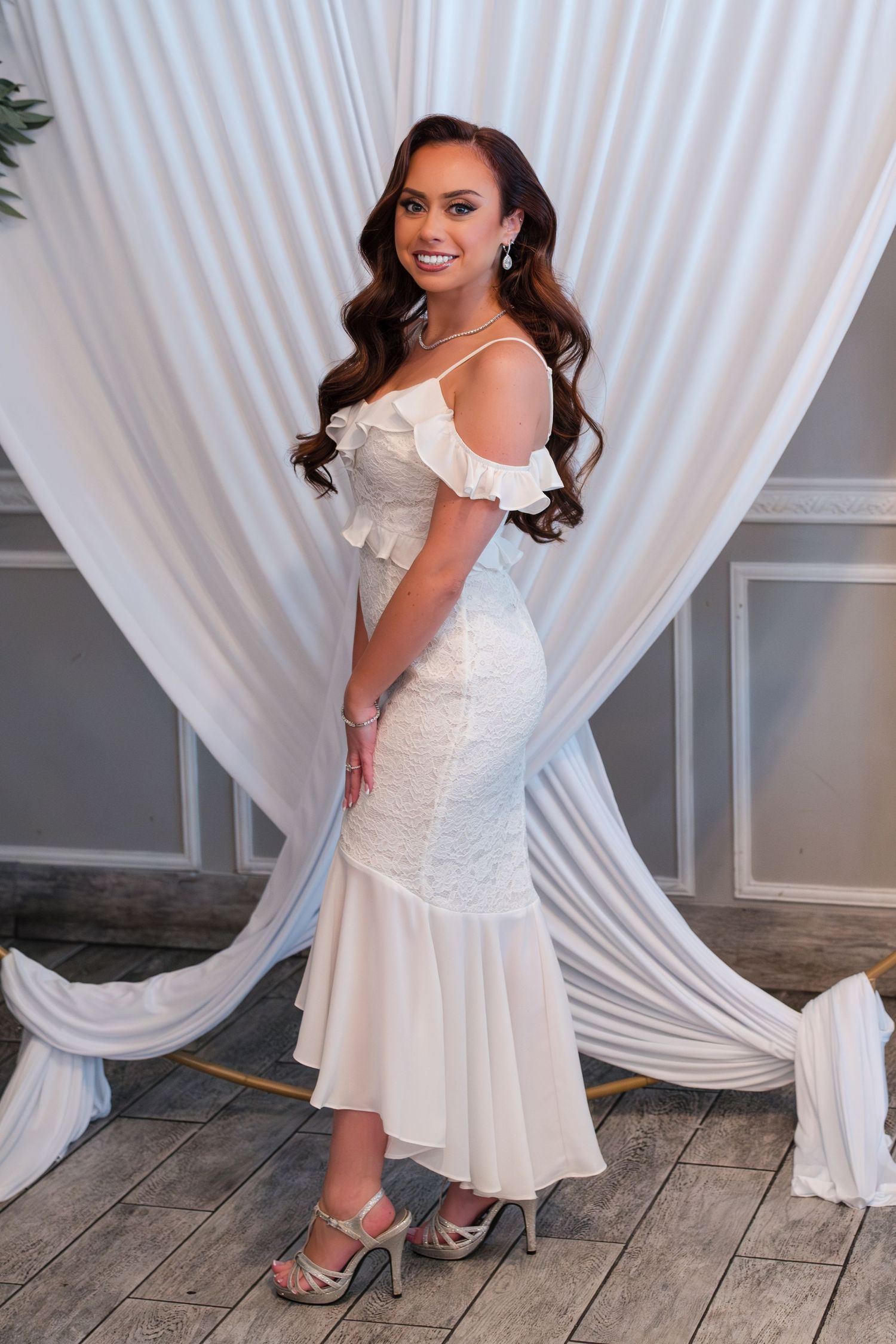 Bride in off-shoulder white mermaid dress posing against draped white backdrop.