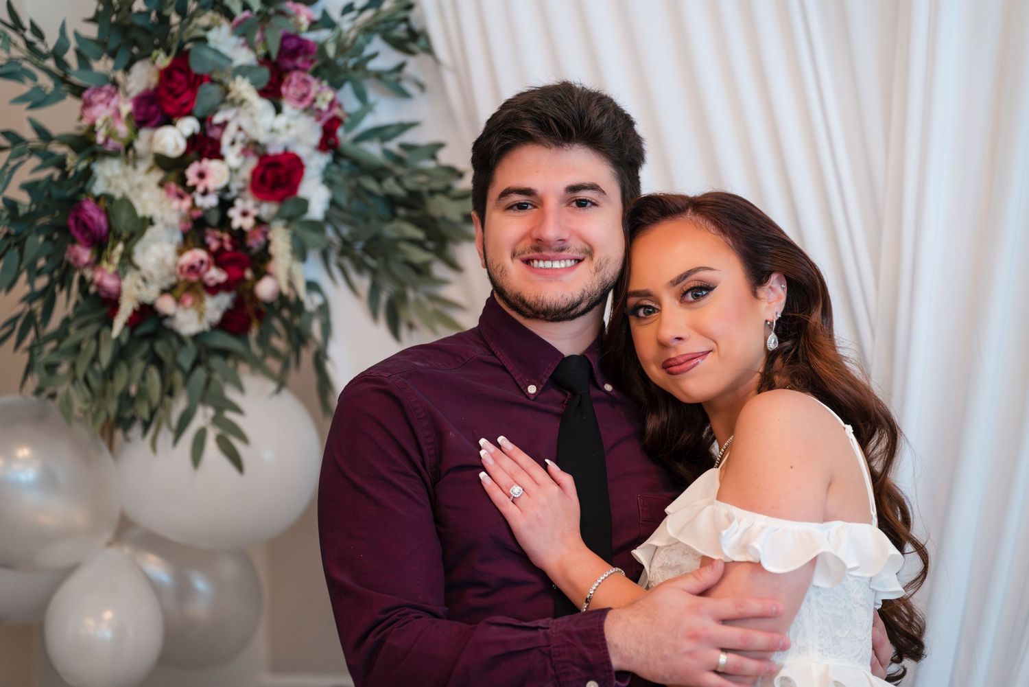 Engaged couple embracing in front of white backdrop with burgundy floral arrangements.