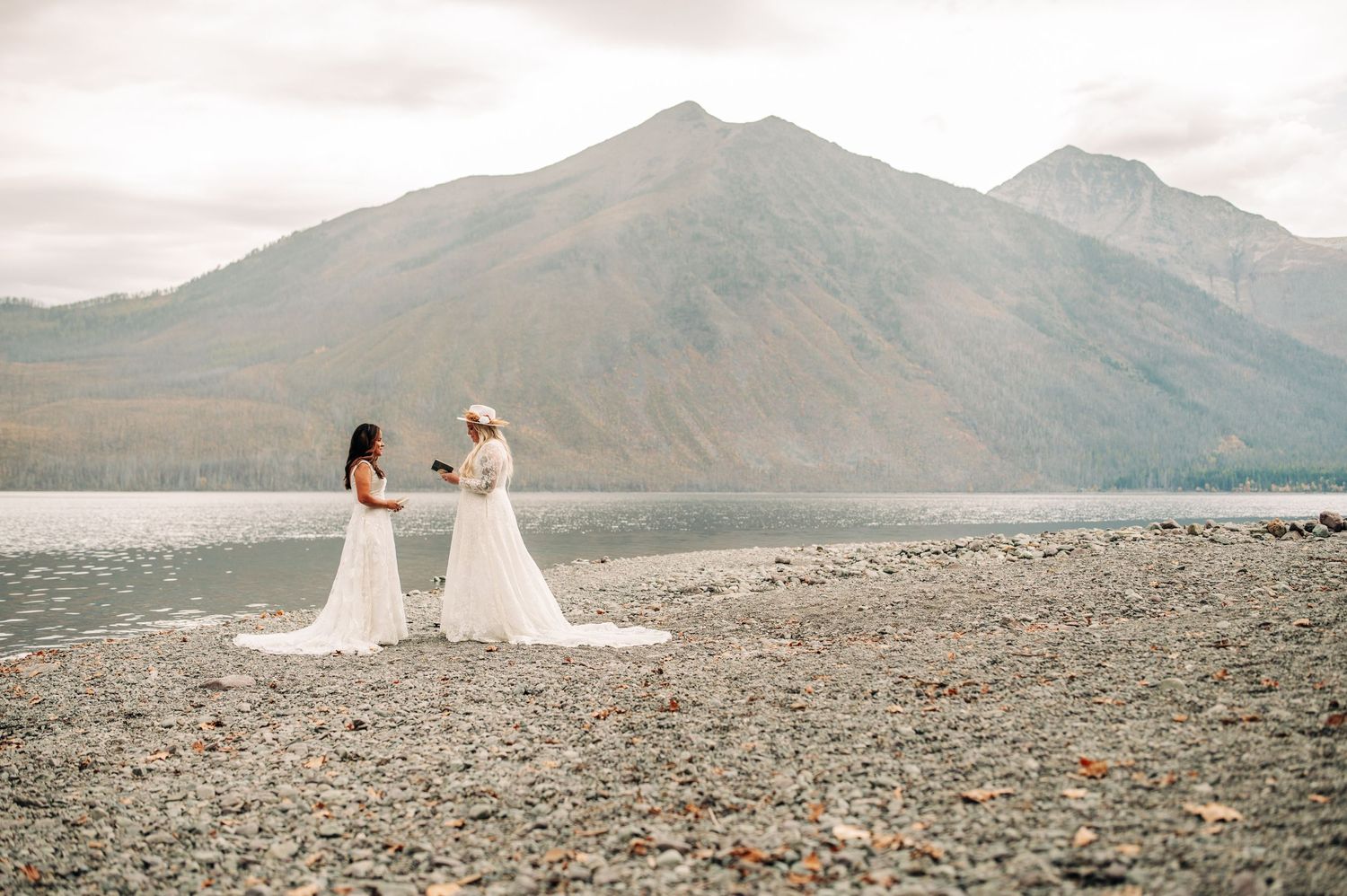Two white wedding dresses stand out against a dramatic mountain lake backdrop on a rocky shore.