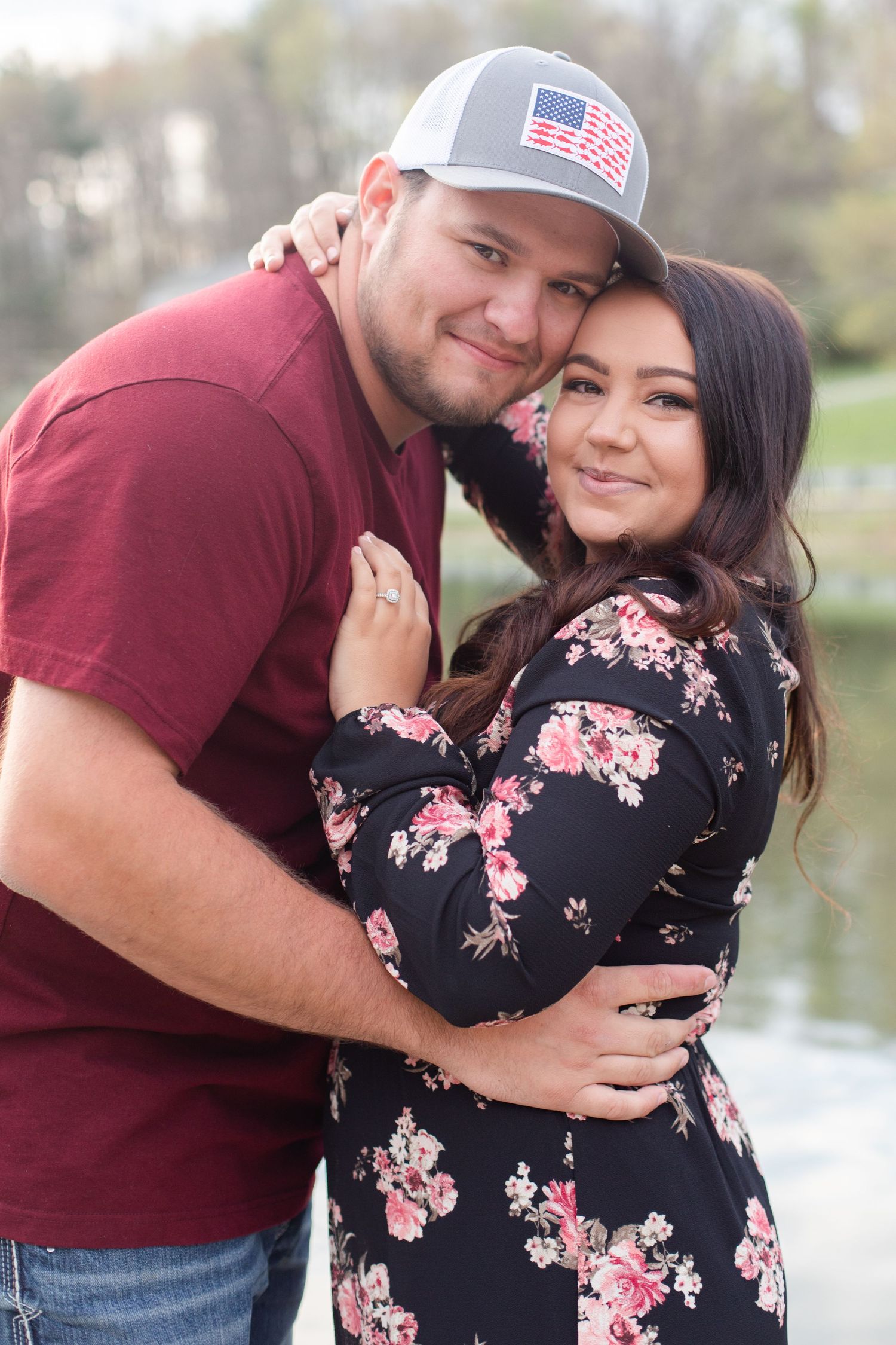 Couple embraces by a lake with her wearing a floral dress and him in a burgundy shirt and American flag cap.