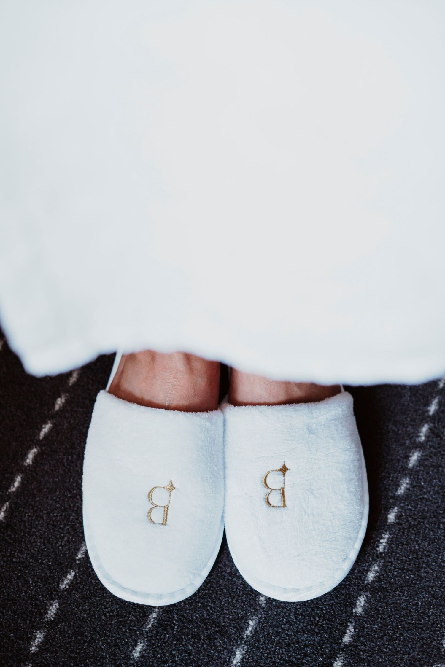 White hotel slippers with embroidered letter B on dark carpet.
