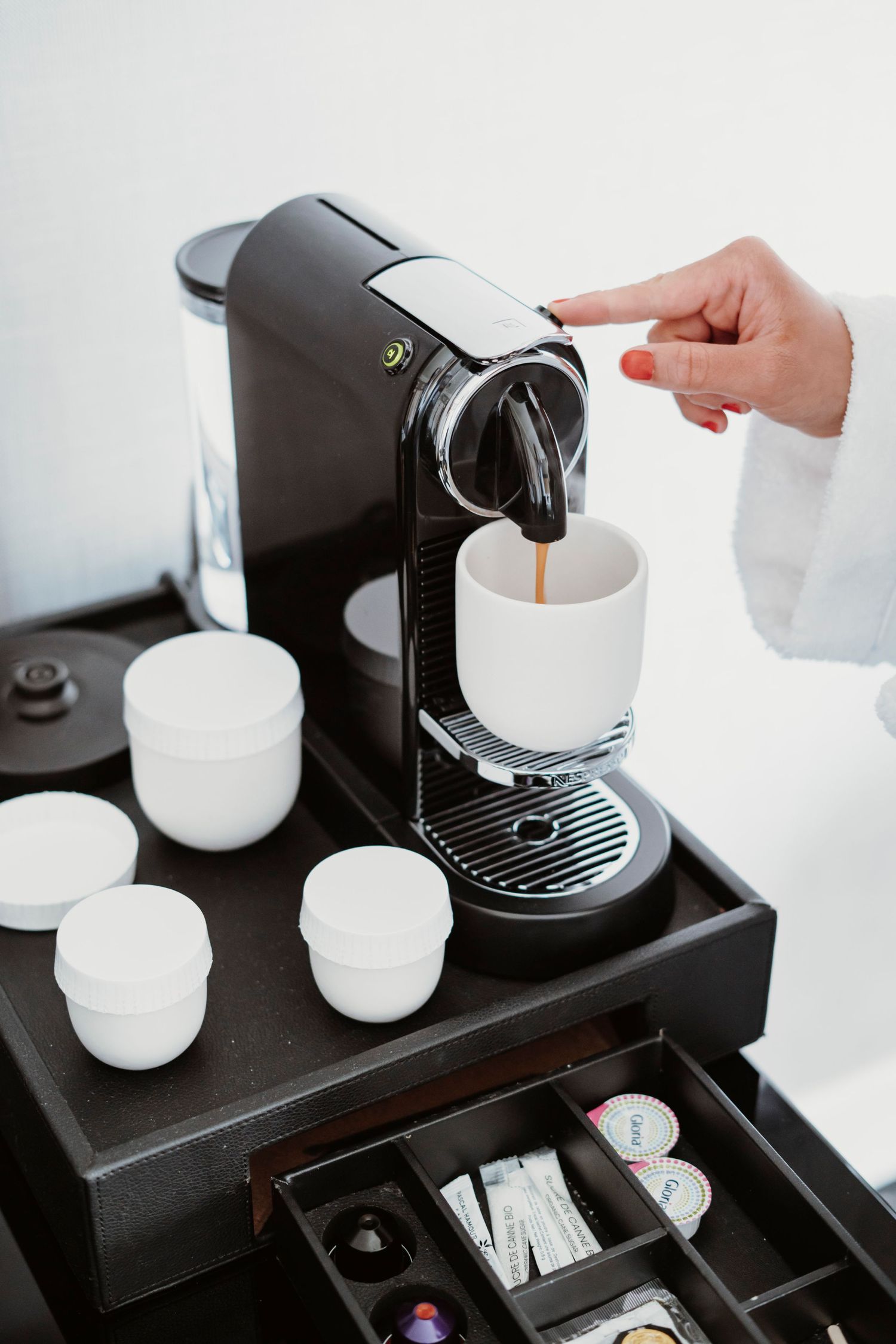 Coffee maker setup with white cups and accessory drawer on dark stand.