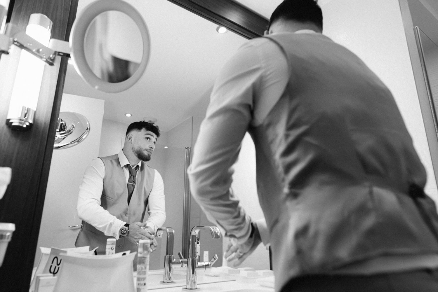 Man in formal attire adjusting his appearance in a bathroom mirror.