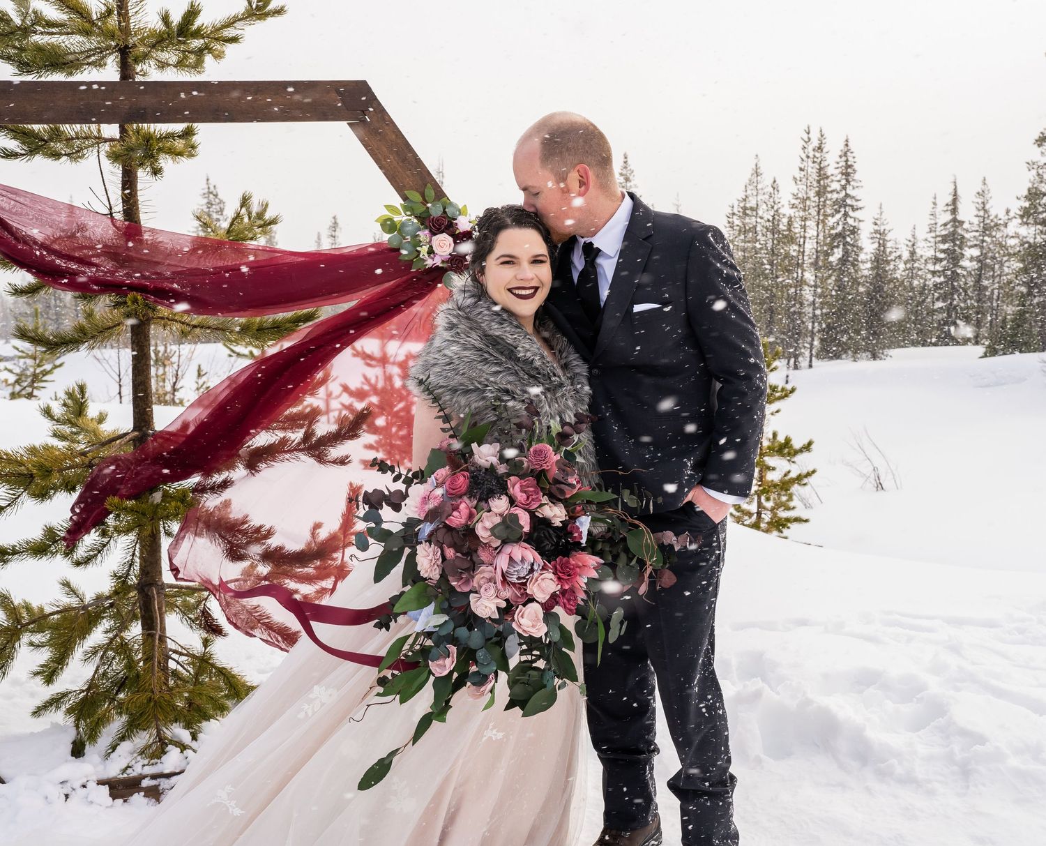 Winter wedding scene with red floral arch and snowy mountain backdrop.