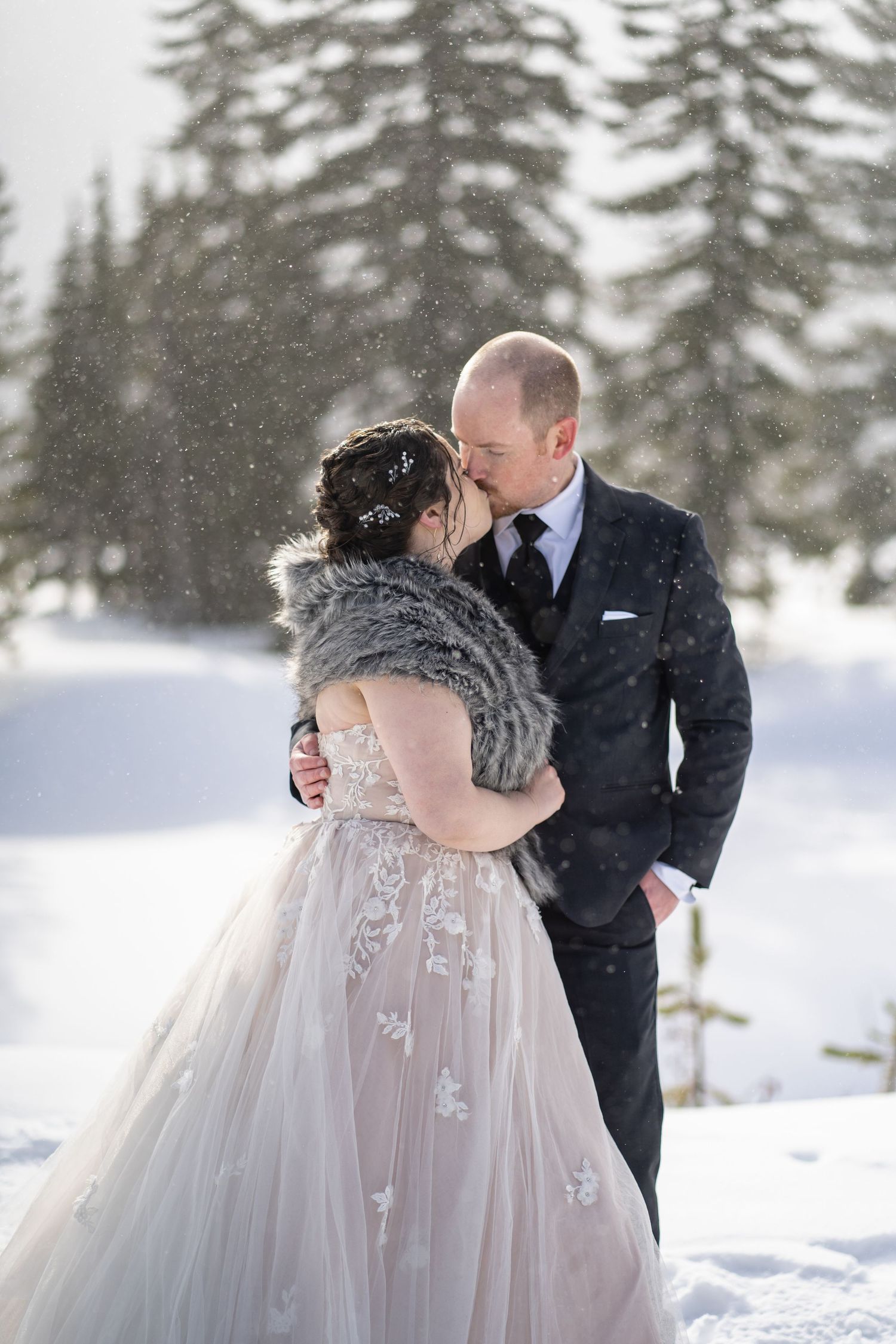 Intimate winter wedding moment with snowfall and evergreen trees in background.
