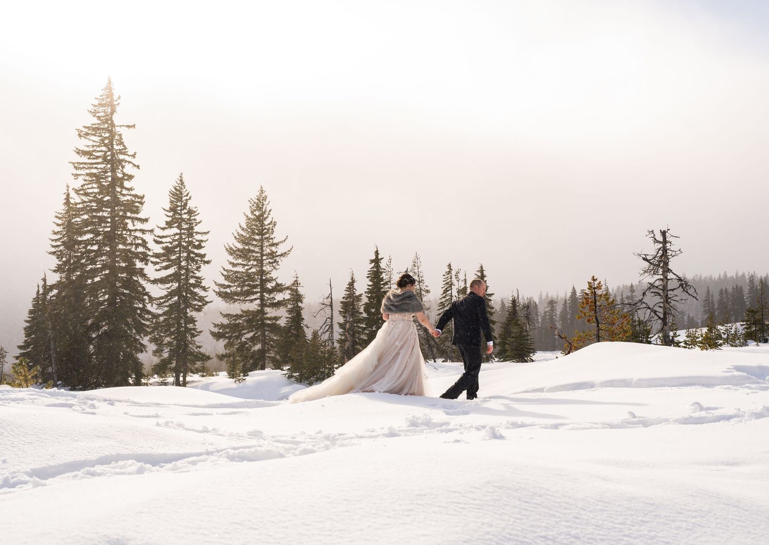 Newlyweds walking through snowy landscape with evergreen trees on misty winter day.